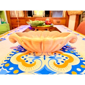 May include: A light peach-colored, scalloped ceramic bowl with curved handles. The bowl is on a table with a patterned tablecloth featuring blue, yellow, and white designs. Other serving dishes are visible in the background.