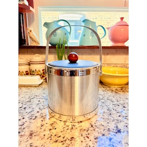 May include: A vintage silver ice bucket with a black lid and a wooden knob. The bucket has a handle and is sitting on a granite countertop. Other kitchenware is visible in the background, including teapots and jars.