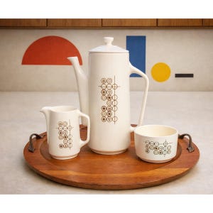 May include: A white ceramic coffee set with a matching creamer and cup, all adorned with a repeating brown geometric pattern. The set sits on a round wooden tray with metal handles, perfect for serving coffee or tea.