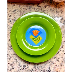 May include: A stack of three green plates. The smallest plate has a central design of a yellow tulip with green leaves on a blue and yellow background. The plates are arranged in a nested fashion, with the largest plate at the bottom.