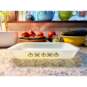 May include: A rectangular, off-white Pyrex baking dish with a floral design in shades of green and gold. The dish is empty and sits on a speckled countertop. Other kitchen items are visible in the background.