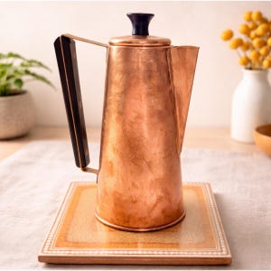 May include: A vintage copper coffee pot with a black handle and lid finial. The pot is placed on a decorative tile. The copper has a warm, reddish-brown hue. The handle is made of dark wood. The pot is approximately 10 inches tall.