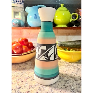 May include: A tall, decorative ceramic vase with a white top and base, and a colorful body. The body features horizontal stripes in teal, peach, and black, with a geometric design. The vase is displayed on a speckled countertop.