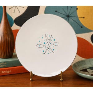 May include: A white ceramic plate with a silver rim, featuring a black and teal abstract design. The plate is displayed on a gold-colored stand, showcasing a mid-century modern aesthetic. A wooden vase and books are visible in the background.