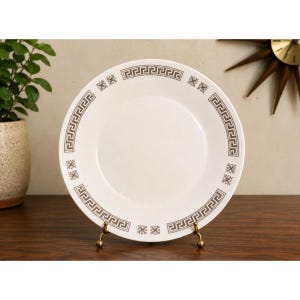 May include: A white ceramic plate with a brown Greek key border and floral accents. The plate is displayed on a gold-colored stand, showcasing its design. The plate is a decorative item.