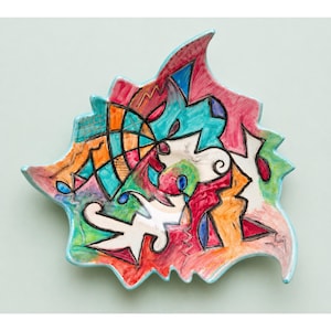 May include: A colorful, abstract ceramic plate with a unique, irregular shape. The plate features a vibrant mix of colors including red, blue, orange, and green, with a light blue rim. The design is reminiscent of modern art.