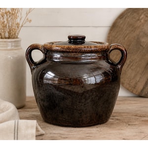 May include: A dark brown ceramic crock pot with a lid and two side handles. The crock pot has a glossy finish and a lighter brown rim. The background includes a wooden surface and a vase with dried plants.
