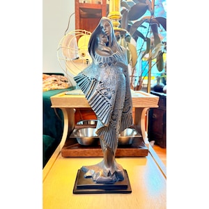 May include: A bronze-colored sculpture of two figures embracing, set on a black rectangular base. The figures are draped in detailed, flowing garments with geometric patterns. The sculpture has a stylized, Art Deco aesthetic.