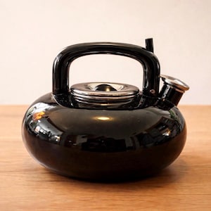 May include: A glossy black kettle with a rounded body, a curved handle, and a silver-colored spout and lid. The kettle sits on a wooden surface, reflecting the light. The design is simple and modern.