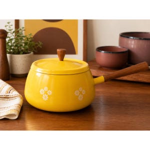 May include: A yellow enamel saucepan with a wooden handle and lid. The saucepan has a floral pattern and a wooden knob on the lid. The handle is attached to the side of the pan. The saucepan is sitting on a wooden surface.