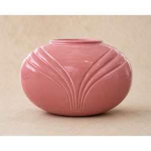 May include: A pink ceramic vase with a rounded, oval shape. The vase features a series of vertical, curved ridges that converge towards the base, creating a stylized design. The vase has a smooth, glossy finish and a wide opening.