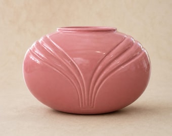 May include: A pink ceramic vase with a rounded, oval shape. The vase features a series of vertical, curved ridges that converge towards the base, creating a stylized design. The vase has a smooth, glossy finish and a wide opening.