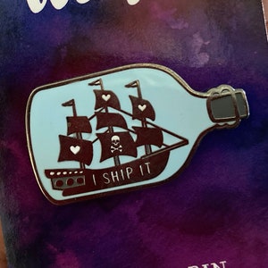 May include: A blue glass bottle with a cork top and a black and white ship inside. The ship has a skull and crossbones and the words "I Ship It" on the sail. The bottle is a pin.