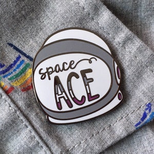 May include: A white enamel pin shaped like an astronaut helmet with the text "space ACE" in black and gray lettering. The "ACE" is in the asexual pride flag colors.