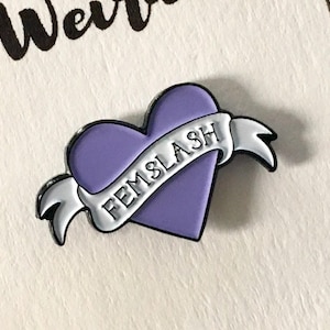 Pode incluir: Pin de esmalte em forma de coração roxo com uma faixa branca que diz "FEMSlash".