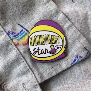 May include: A purple and yellow enamel pin with a space helmet design. The helmet has a white background with the text "nonbinary star" and a small planet with a star on it.