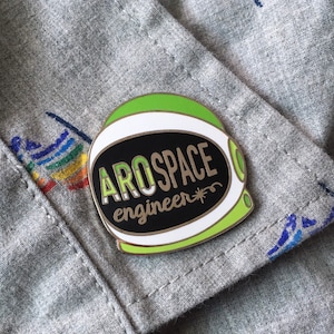 May include: A green and white enamel pin shaped like an astronaut helmet with the text "Aerospace engineer" in gold lettering on a black background.