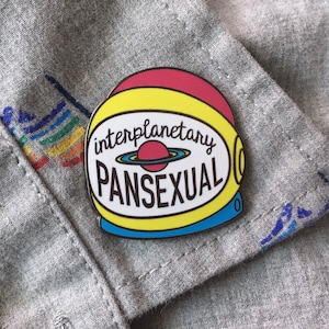 May include: A yellow, red and blue enamel pin shaped like an astronaut helmet with the text "interplanetary pansexual" inside.