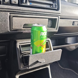 May include: A green can of Rockstar Energy drink is placed in the cup holder of a car's centre console. The can is partially visible, with the Rockstar logo and the words "Energy Drink" visible.