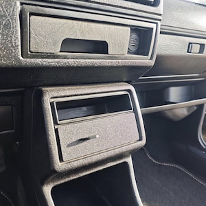 May include: Close-up of the interior of a car, showing a black dashboard with a storage compartment and a cigarette lighter.
