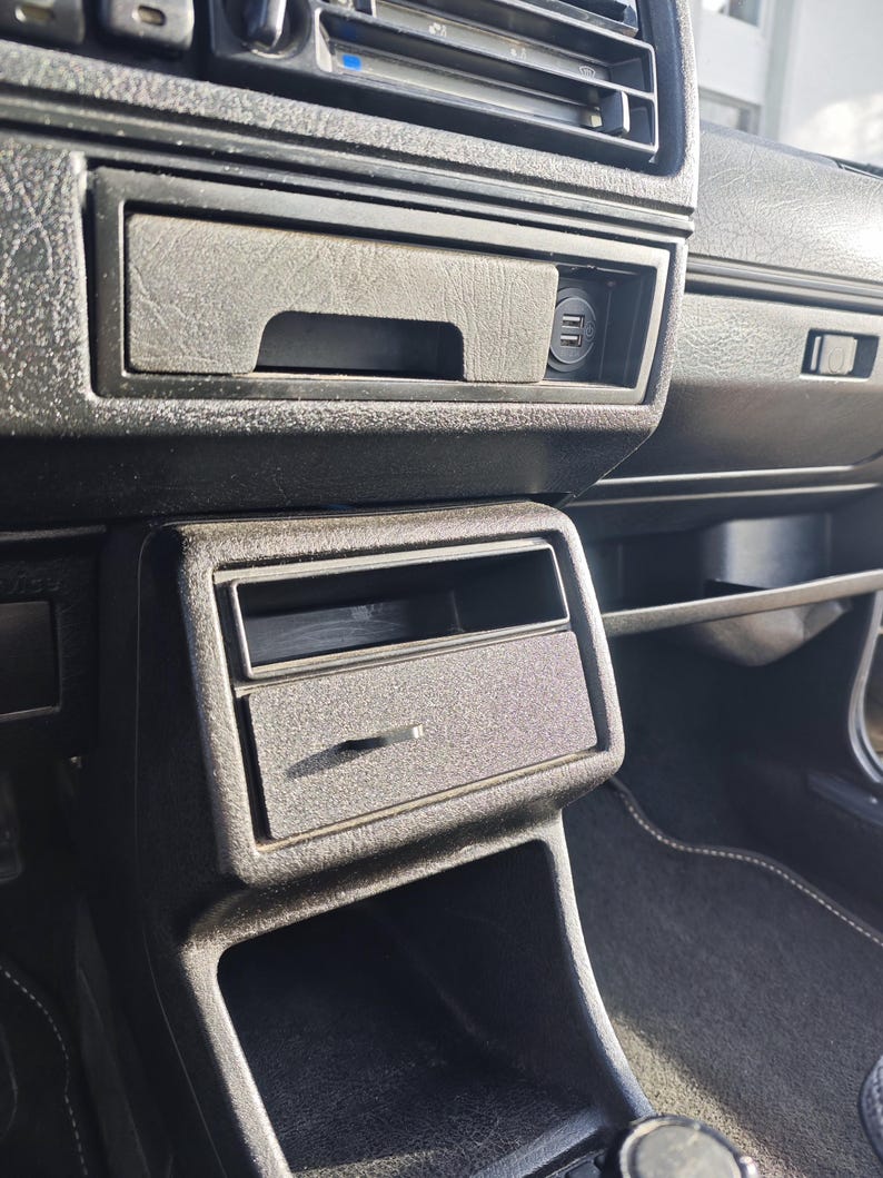 May include: Close-up of the interior of a car, showing a black dashboard with a storage compartment and a cigarette lighter.