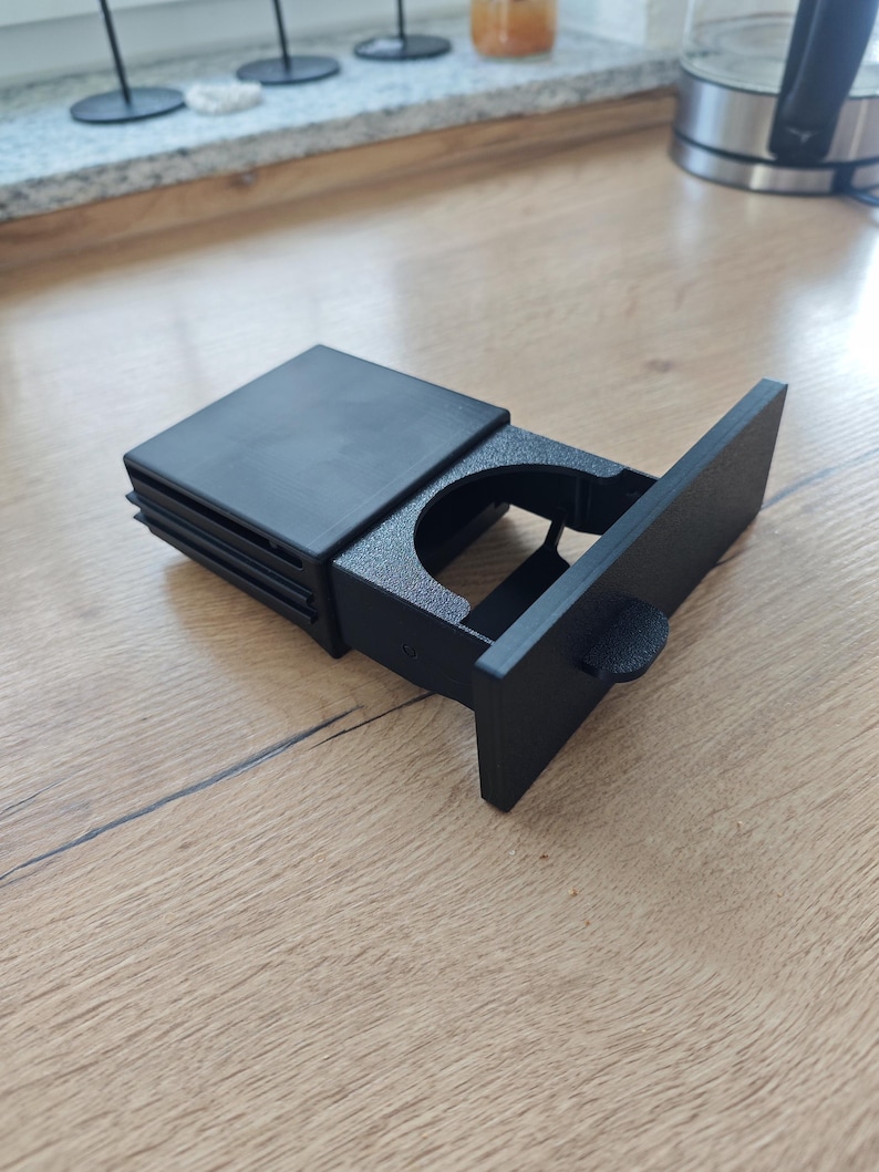 May include: Black plastic cup holder with a sliding lid. The cup holder is designed to fit in a car's centre console.