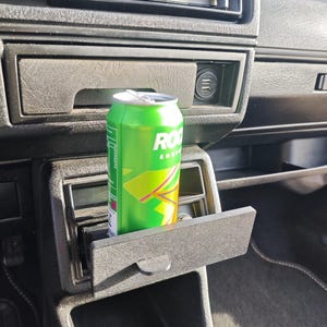 May include: A green can of Rockstar Energy Drink is placed in a black car cup holder. The cup holder is located in the centre console of the car.