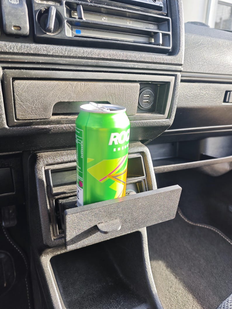 May include: A green can of Rockstar Energy Drink is placed in a black car cup holder. The cup holder is located in the centre console of the car.