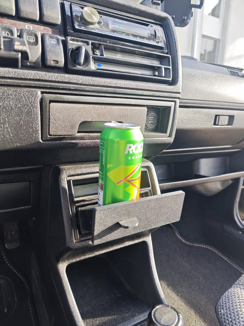 May include: A green can of Rockstar Energy drink is placed in the cup holder of a car's centre console. The can is partially visible, with the Rockstar logo and the words "Energy Drink" visible.