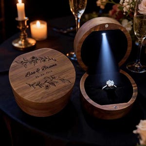 May include: Two wooden ring boxes, one closed and one open, with a diamond ring illuminated by a spotlight. The closed box has the names "Lisa & Briana" and the date "06-09-2026" engraved on the lid. The boxes are on a black surface.