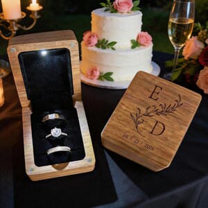 May include: A wooden ring box with a hinged lid, featuring an engraved floral design with the letters E and D and the date 08 22 2026. The open box has a black velvet interior holding three silver rings, illuminated by a small built-in LED light.
