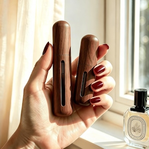 May include: Two cylindrical perfume atomisers with a dark brown wood grain finish, each featuring a vertical transparent viewing window. The atomisers are held together, with one being taller than the other, showcasing a smooth, rounded top design.
