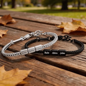 May include: Two braided chain bracelets with square beads. One bracelet is silver-toned with beads engraved with the names Gina, Amber, and Calvin. The other bracelet is black with beads engraved with the names Rain, Abira, and John. Both feature metal clasps.