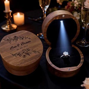 May include: A round wooden ring box with a dark wood grain finish. The lid is engraved with floral branches and the text Lisa & Briana 06-09-2026. An open matching box features a black interior, a built-in LED light, and a silver diamond ring.