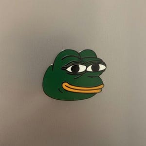 Op de afbeelding: Een groene, kikkerachtige cartoonfiguur met grote, verdrietige ogen en een gele mond. De figuur is een populair internetmeme, tegen een grijze achtergrond.