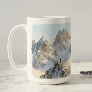 Puede incluir: Taza de cerámica blanca con un diseño de paisaje montañoso. La imagen muestra a una persona en la cima de una montaña nevada. La taza tiene un asa grande y un acabado brillante, ideal para café o té.