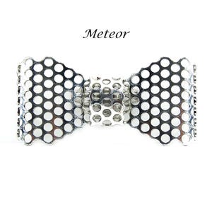 May include: A silver-toned metal bow-shaped accessory featuring a perforated grid pattern with circular cutouts throughout the entire surface. The word Meteor is printed in black text above the bow, which has a central band and scalloped edges.
