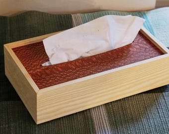 May include: A rectangular wooden tissue box with a light-colored frame and a reddish-brown textured top. White tissues are partially pulled from the opening. The box is placed on a patterned surface.