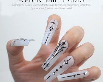 Pode incluir: Close-up de uma mão com unhas compridas, brancas, em forma de amêndoa com desenhos geométricos em preto e branco e strass. As unhas são decoradas com um padrão de estrela.