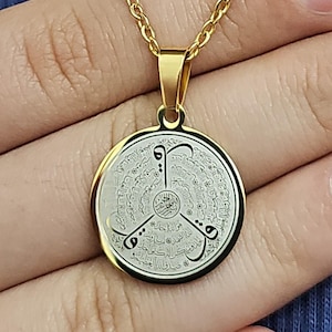 May include: Gold chain necklace with a round pendant featuring Arabic calligraphy in black and white on a white background.