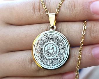 Può includere: Collana con pendente dorato con un pendente circolare con intricata calligrafia bianca e nera. Il pendente è sospeso a una delicata catena d'oro. Il testo "Ayetel Kürsi" è visibile sul pendente.
