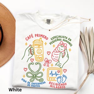 May include: White t-shirt with colorful illustrations and text. The design includes a coffee cup, a baby, a stethoscope, and a heart monitor. Text includes "CAFE PRIMERO", "ESPECIALISTA EN PEQUEÑOS HUMANOS", "VIDA DE ENFERMERA DE PARTOS", and "RECIBIENDO MILAGROS".