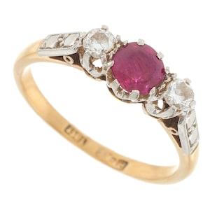 Puede incluir: Un anillo de oro con un rubí rojo y dos diamantes. El anillo está engastado en un diseño delicado y ornamentado.