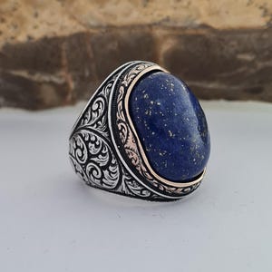 Könnte beinhalten: Ein silberner Ring mit einem ovalen Lapislazuli-Stein, der in einer dekorativen Silber- und Roségold-Banderole sitzt.