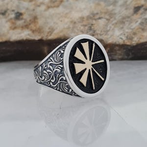 May include: A silver ring with an oval face featuring a black background and a gold-colored starburst design. The band is detailed with an ornate, swirling pattern. The ring is displayed on a reflective surface.