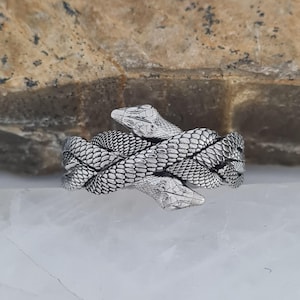 May include: A silver-toned metal ring featuring a detailed design of two intertwined snakes. The snakes' bodies are textured with a realistic scale pattern, and their heads are positioned to face opposite directions, overlapping at the centre.