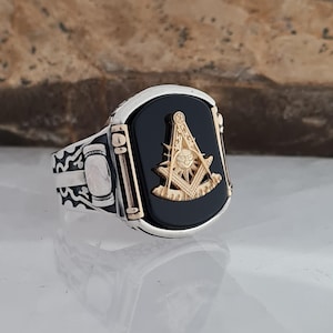 Pode incluir: Anel prateado com uma pedra central de ônix preto. O anel apresenta um símbolo maçónico dourado. A banda tem padrões decorativos e um design retangular. O anel é exibido numa superfície refletora.