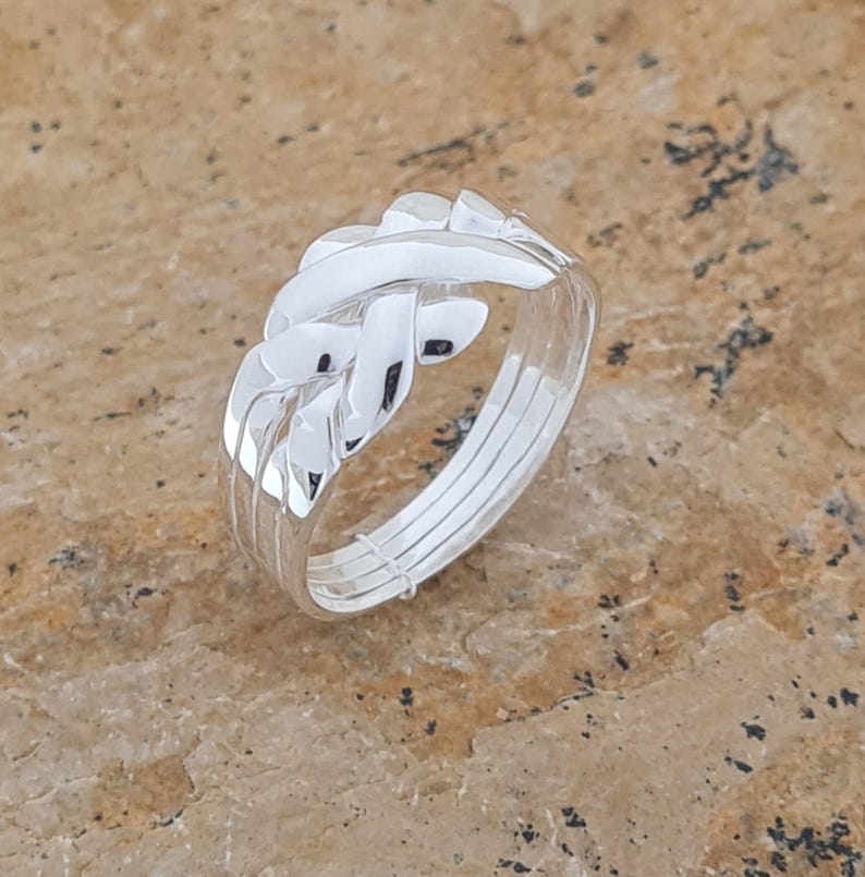 May include: A silver puzzle ring with a woven design. The ring has a complex, interlocking pattern, with multiple bands forming the ring's structure. The metal has a polished, reflective surface, and the ring is set against a neutral background.