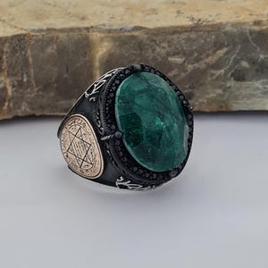 May include: A silver ring featuring a large green gemstone set within a black stone surround. The ring has an elaborate design with Arabic script on the side.