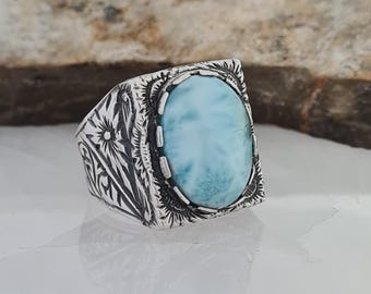 May include: A silver ring featuring a light blue oval gemstone. The ring has an engraved pattern around the stone and on the band. The gemstone is set in a bezel setting. The ring is a statement piece, suitable for everyday wear.